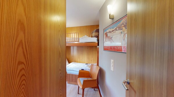 A compact room with a bunk bed, a small desk, and a chair. A poster on the wall depicts a winter scene.