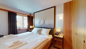 A hotel room with a large bed, two bedside lamps, a window with curtains, and a wall-mounted artwork of deer. - 3-room apartment on the first floor, 67m2. (Celerina/Schlarigna)