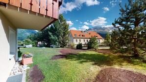 Ein Haus mit rotem Dach, ein Garten mit Rasen und Bäumen, und Berge im Hintergrund.