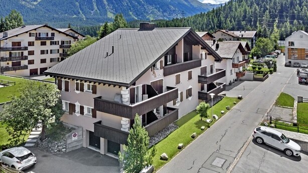 A modern multi-story building with balconies, surrounded by a residential area and mountains in the background. - 2-room apartment on the 1st floor. (Celerina/Schlarigna)