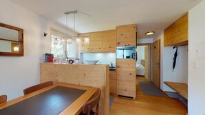 A compact kitchen with wooden cabinets, a dining area with a table and chairs, and a visible kitchen counter with various items. - 2-room apartment on the 1st floor. (Celerina/Schlarigna)