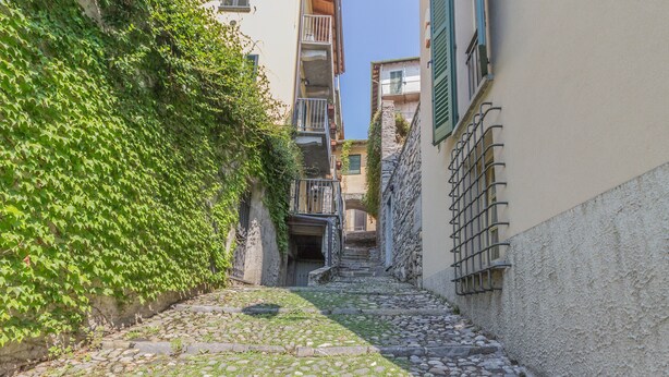 Une ruelle étroite pavée de pierres, bordée de bâtiments en pierre, avec des balcons et un escalier menant à l’étage supérieur.
