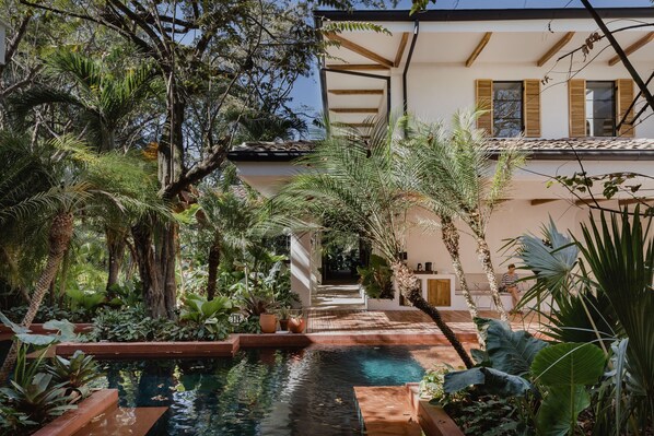 Una piscina rodeada de exuberante vegetación tropical y un edificio con vigas y contraventanas de madera.