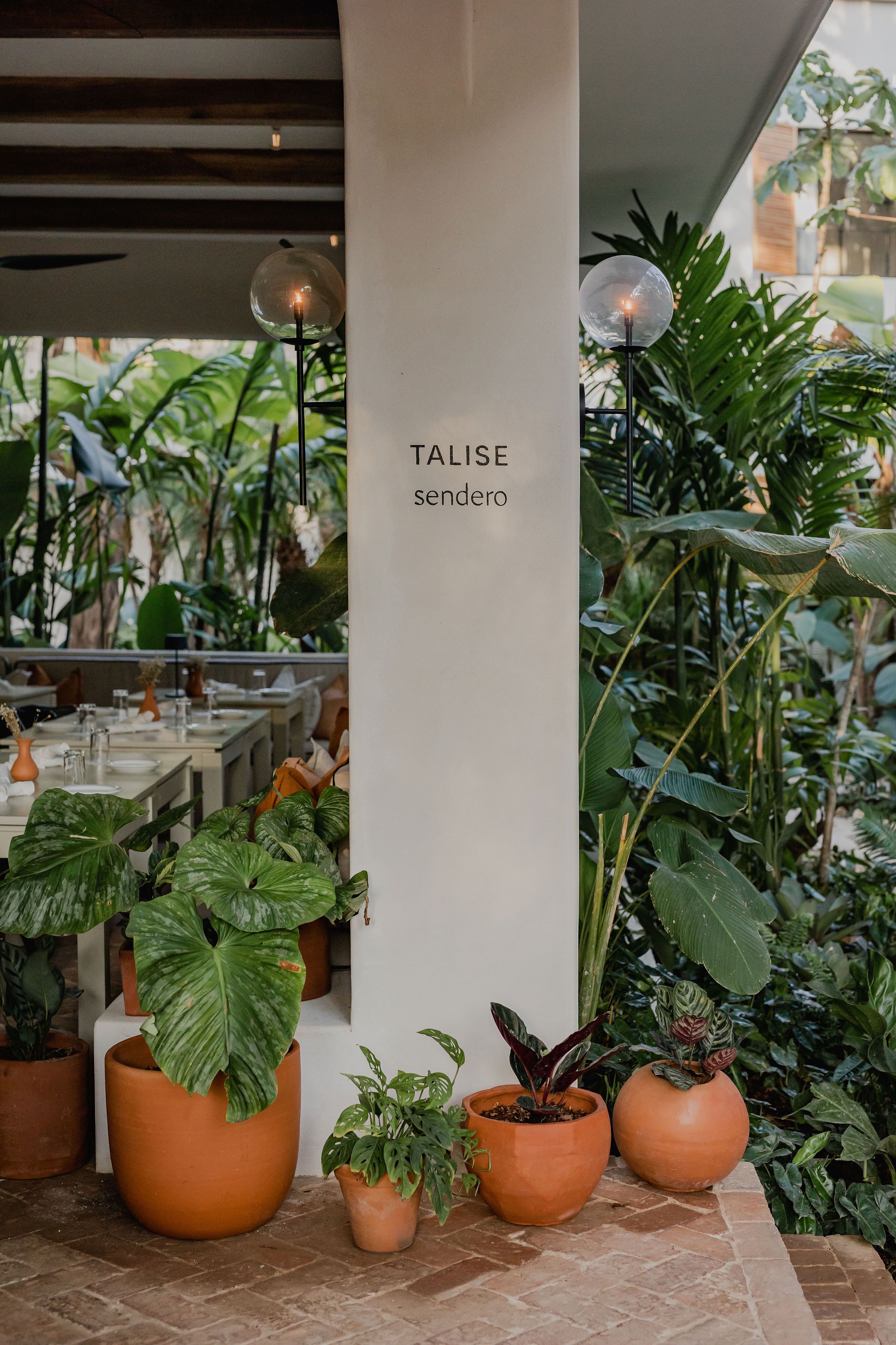 Un ristorante con vasi in terracotta pieni di piante diverse, un pilastro bianco con la scritta 'TALISE sendero' e un pavimento in mattoni.