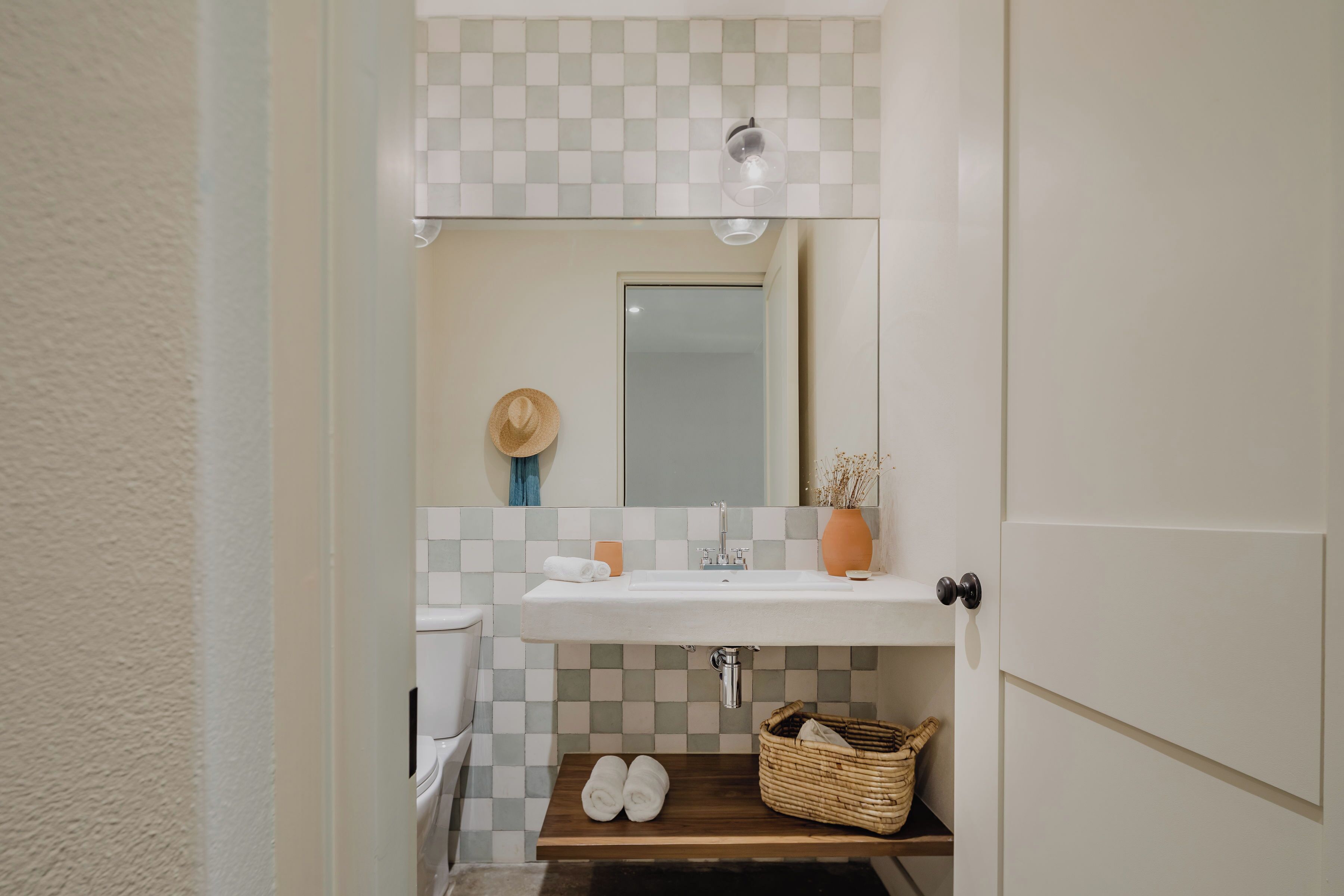 Un baño con salpicadero de baldosas a cuadros, un lavamanos blanco, un espejo, una estantería de madera con toallas y una cesta de mimbre.