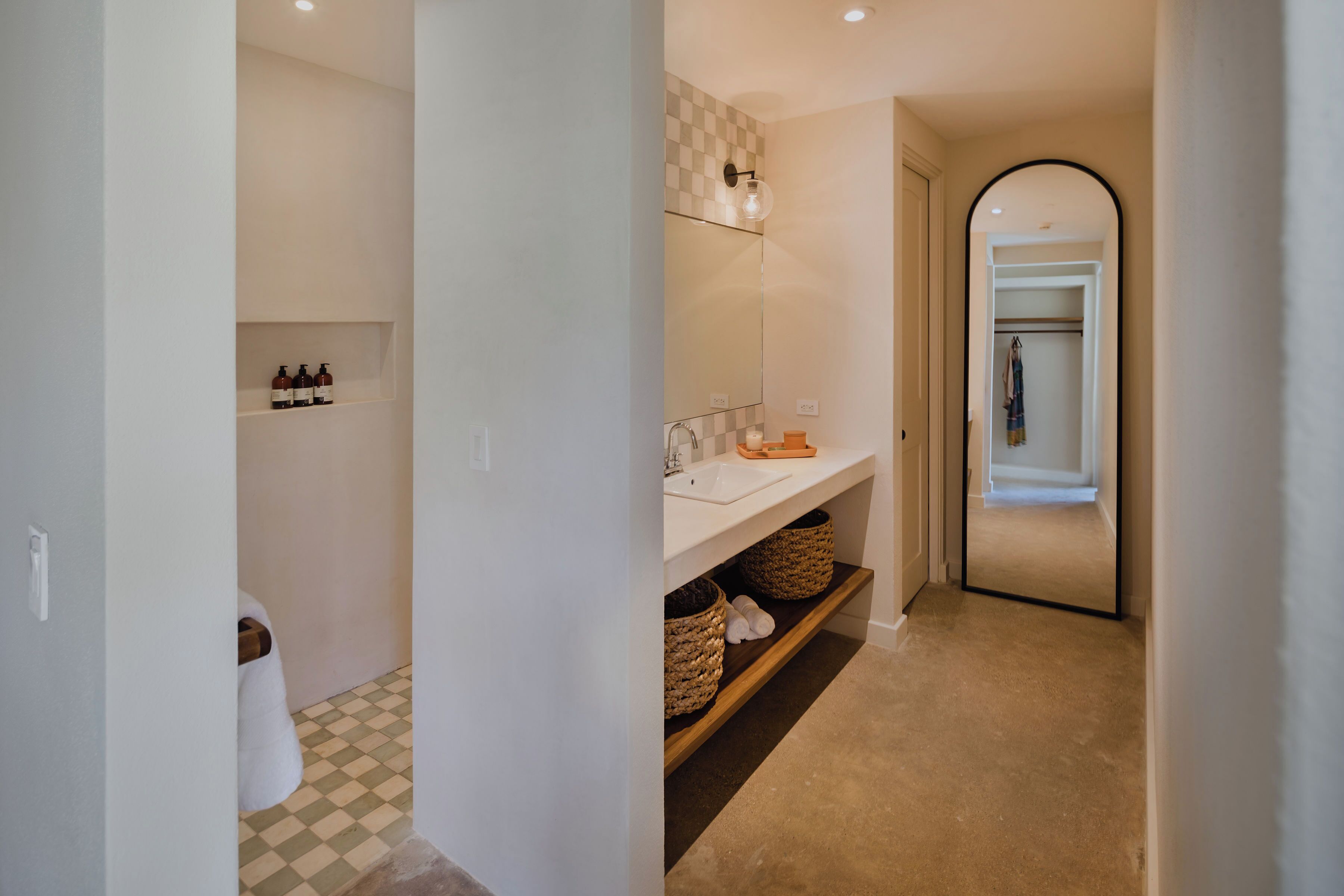 Un pasillo con un baño que tiene piso a cuadros, un lavamanos y un espejo. Hay cestas en una estantería y un perchero para toallas.