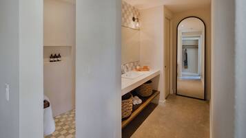 Un pasillo con un baño que tiene piso a cuadros, un lavamanos y un espejo. Hay cestas en una estantería y un perchero para toallas.