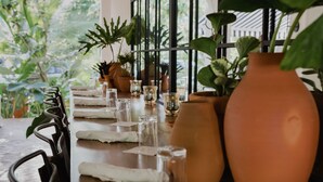 Une table à manger dressée avec des verres, des assiettes et des serviettes en tissu, entourée de plantes en pot et d’un grand pot en terre cuite.
