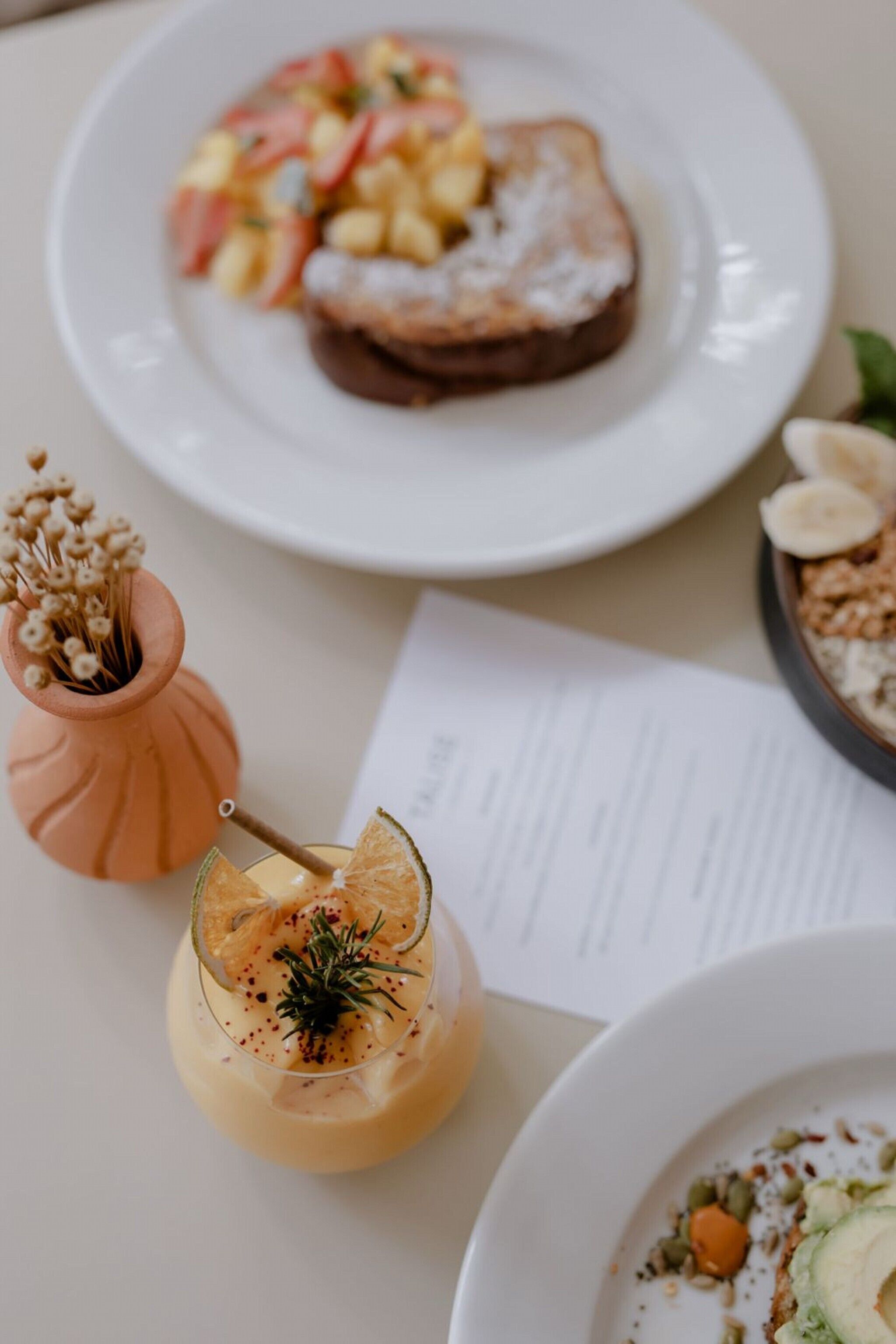 Une table avec une assiette de toasts garnis de fruits, un verre d’une boisson crémeuse décorée d’une branche de romarin, et un bol de granola avec des rondelles de banane.