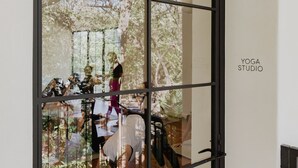 Une porte en verre qui reflète l’intérieur d’un studio de yoga, avec notamment une personne en position de yoga, un tapis de yoga et un arbre à l’extérieur.