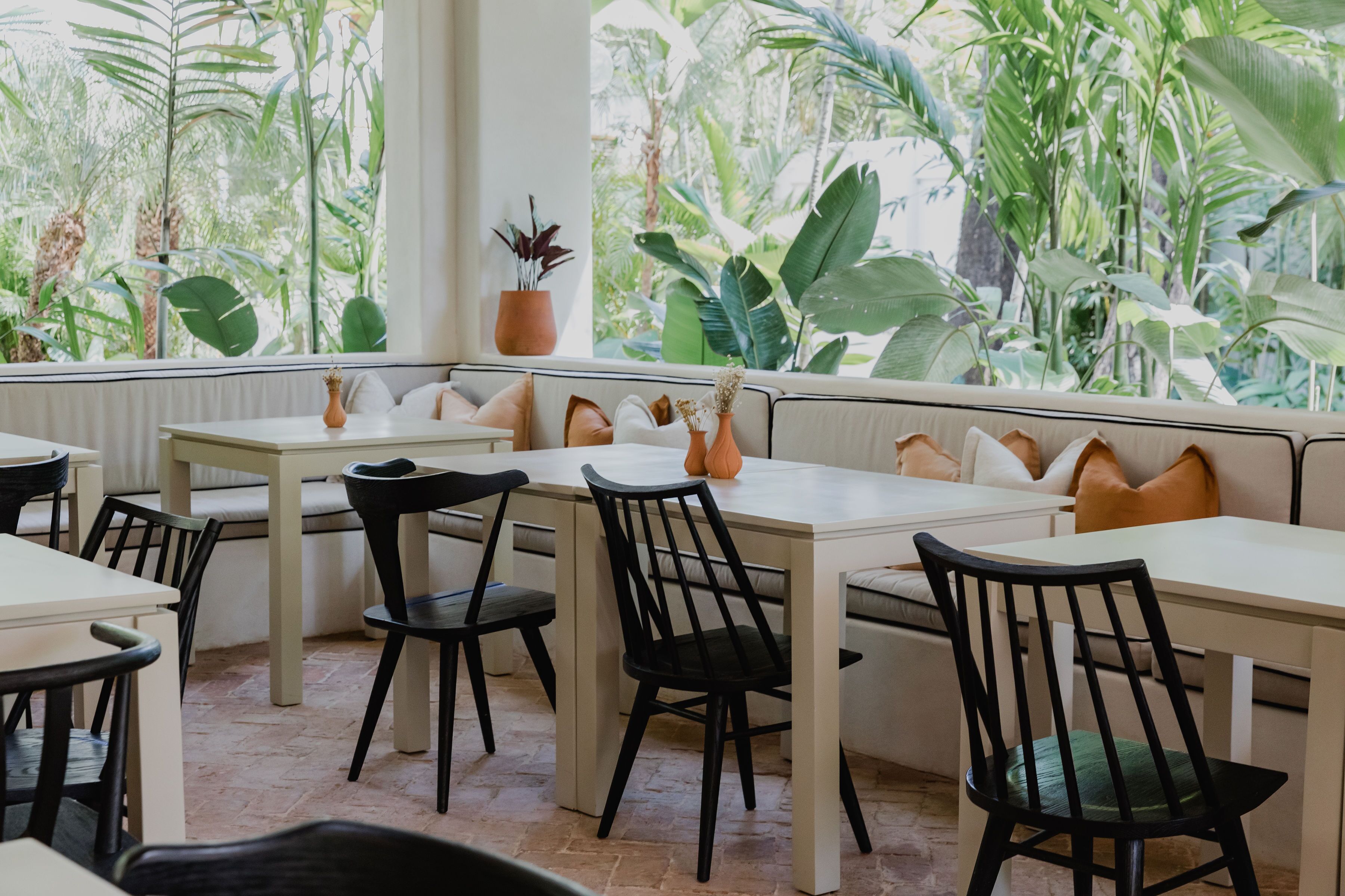 Un restaurante con mesas blancas y sillas negras, amplias ventanas con plantas tropicales y una zona de estar acogedora con cojines.