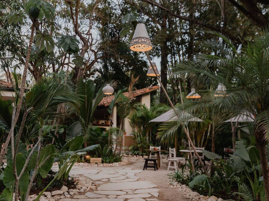Un sentier de jardin mène à un espace aménagé avec des tables et des chaises, entouré d’une végétation tropicale luxuriante et agrémenté de lampes suspendues.