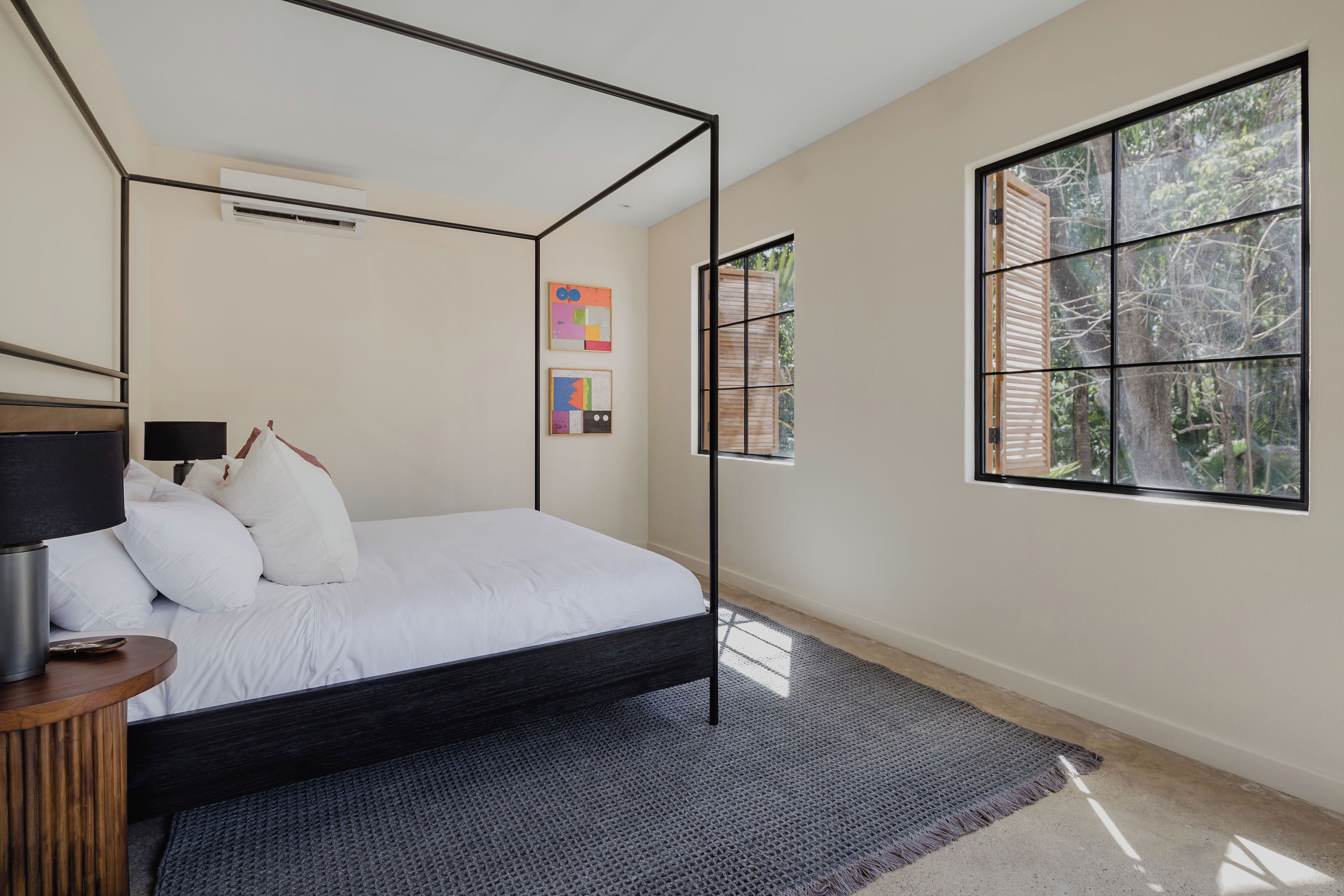 A modern bedroom with a four-poster bed, a bedside table with a lamp, and a large window with a view of trees.