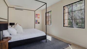 A modern bedroom with a four-poster bed, a bedside table with a lamp, and a large window with a view of trees.