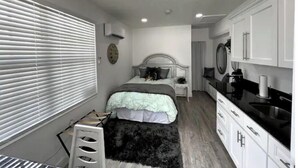A bedroom with a bed, bedside table, and a small desk with a chair. There is a window with blinds, a mirror, and a kitchen area with cabinets and a sink. - Modern studio apartment with covered outside seating area (Spring Hill)