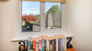 Una habitación con ventana que da a un árbol y un edificio, una estantería con libros y un reloj en la pared.