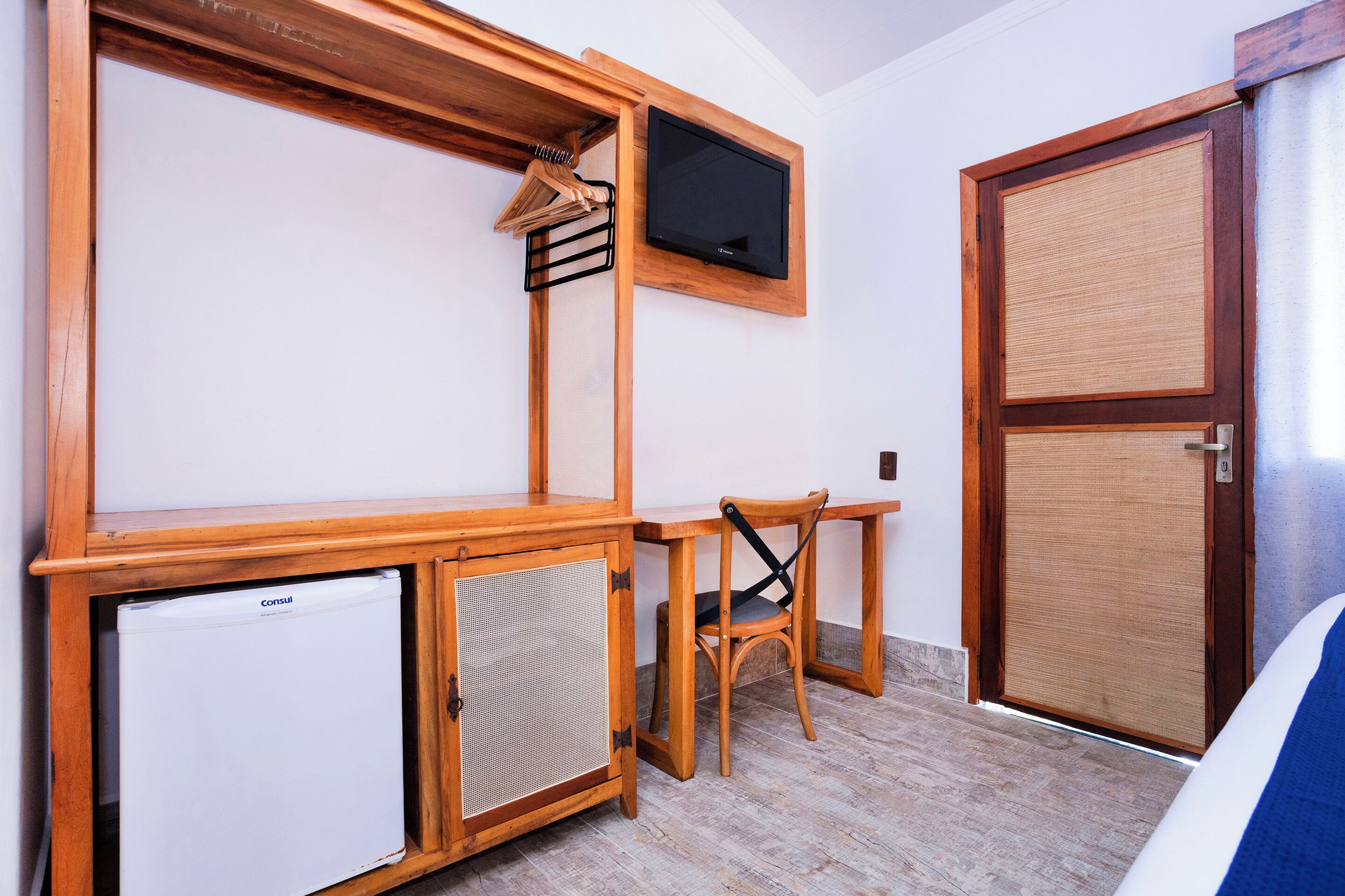Quarto compacto com escrivaninha de madeira, pequena geladeira, cadeira e televisão fixada na parede.