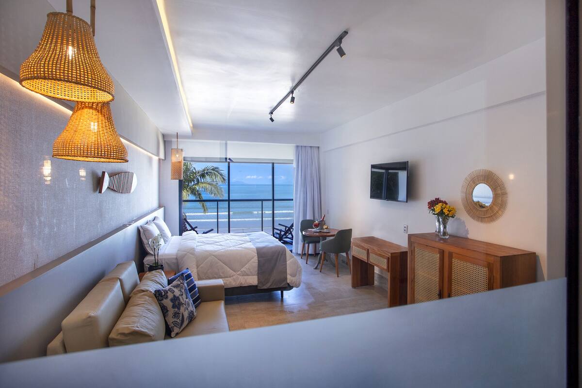 Habitación de hotel moderna con cama, zona de estar, escritorio y un amplio ventanal con vistas al océano.