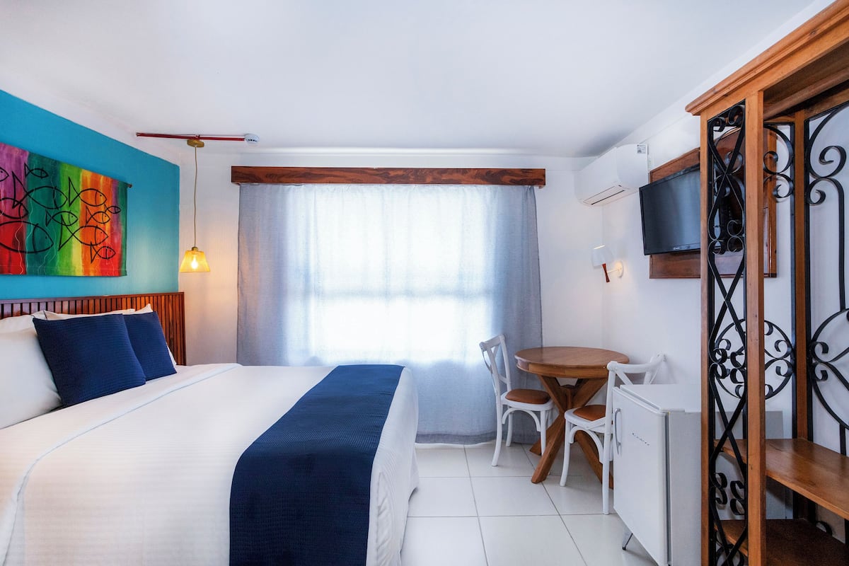 Habitación de hotel con cama, una mesa de comedor con sillas, televisor y un cuadro abstracto y colorido en la pared.