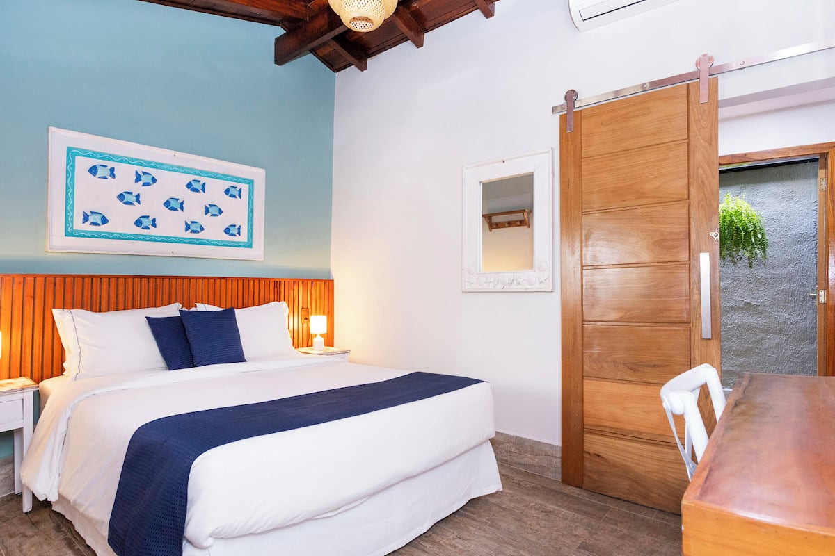 Un dormitorio con cama, mesitas de noche, una puerta de madera, un espejo y un cuadro enmarcado en la pared.