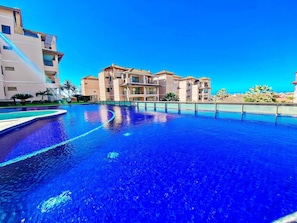 A swimming pool with a clear blue water surface, surrounded by multi-story buildings with balconies and a clear sky.