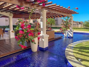 Zona de piscina con una pérgola de madera, plantas en macetas y una piscina azul cristalina.