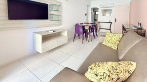 Un salon moderne avec un téléviseur à écran plat, un canapé, une table à manger avec des chaises, et un coin cuisine à l’arrière-plan.