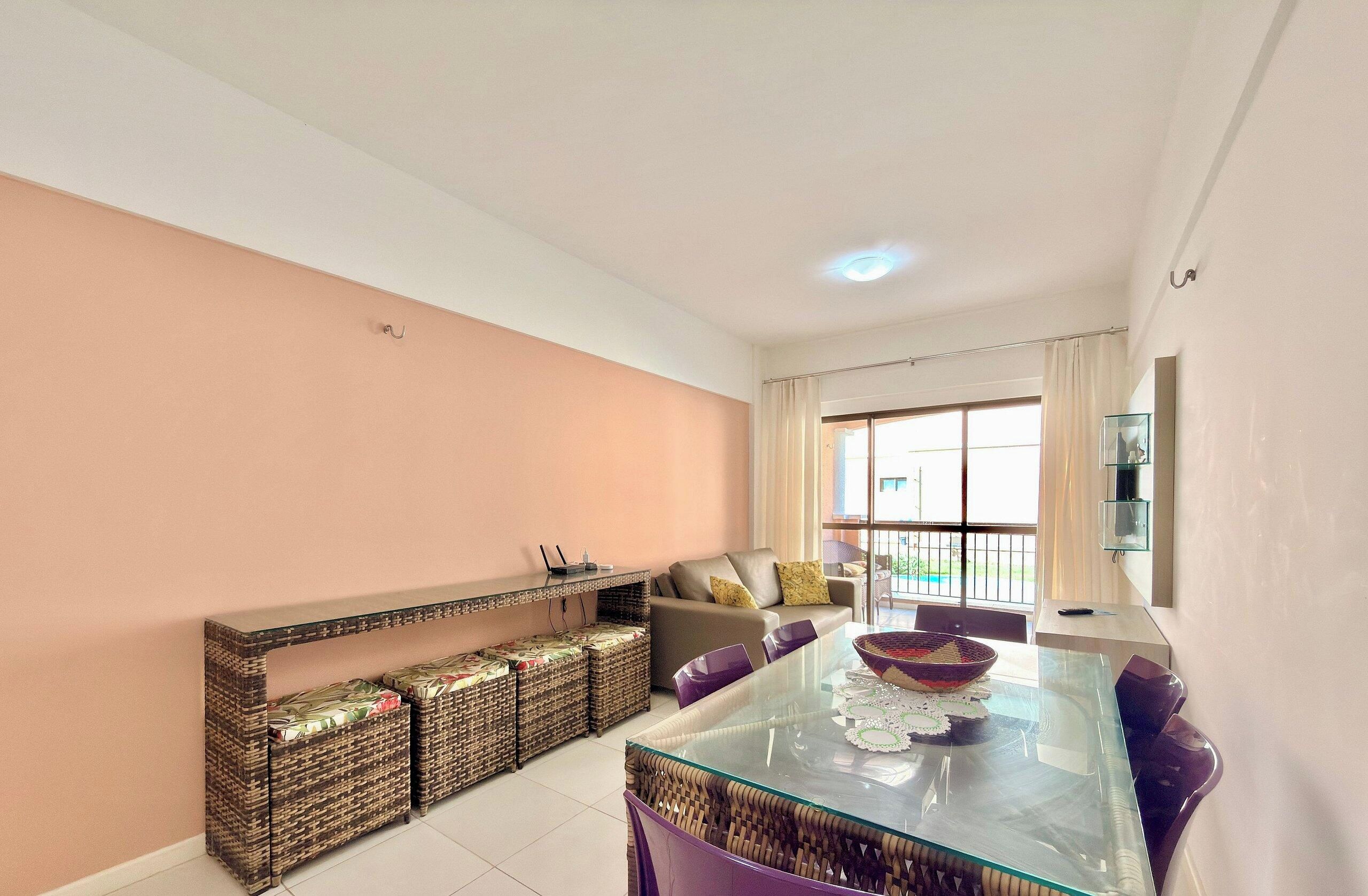 A modern living room with a glass dining table, wicker furniture, a sofa, and a large window with curtains.