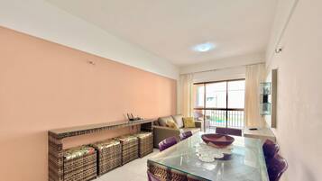 A modern living room with a glass dining table, wicker furniture, a sofa, and a large window with curtains.