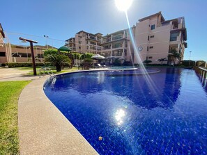 Une piscine à l’eau d’un bleu limpide, entourée d’une promenade dallée et jouxtant un immeuble résidentiel à plusieurs étages doté de balcons.