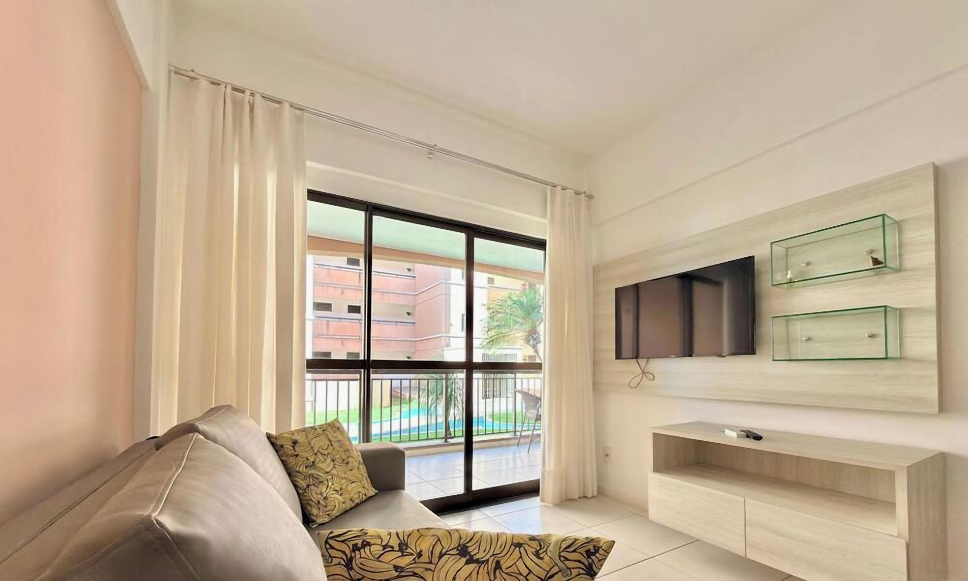 Un salon moderne avec un canapé, un téléviseur à écran plat fixé au mur et un balcon offrant une vue sur les immeubles.