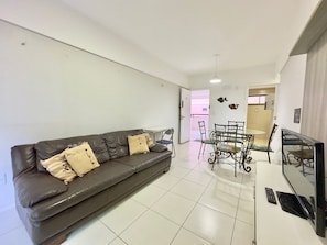 Uma sala de estar com um sofá marrom escuro, uma mesa de jantar com cadeiras, uma televisão e uma cozinha visível através de uma porta aberta.