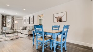 Un coin repas avec une table en bois et des chaises bleues, un canapé et une œuvre d’art sur le thème des taureaux accrochée au mur.