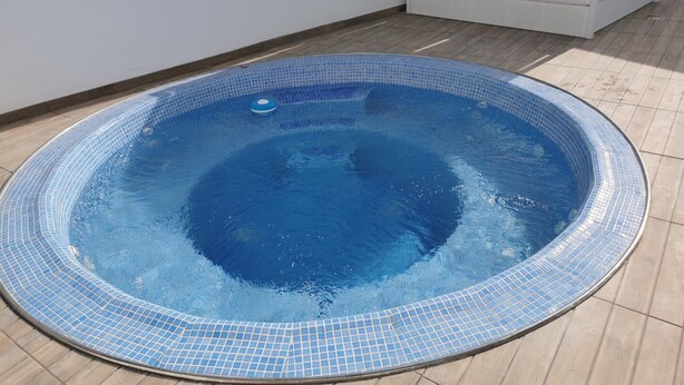 Una piscina rodona amb un videt de rajoles, una cadira de relaxació blanca i un llit blanc al fons.