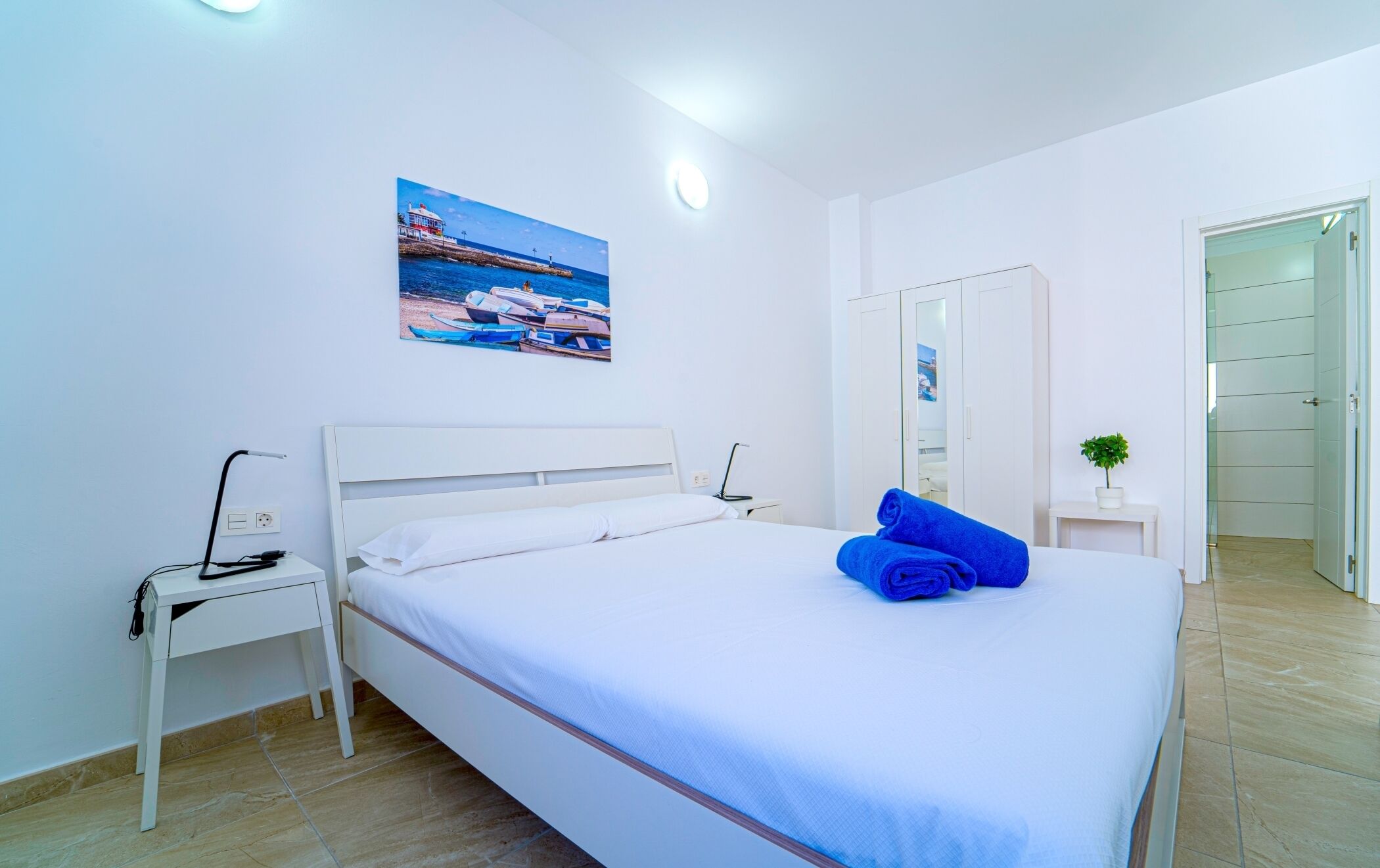 A hotel room with a bed, a nightstand, a desk, a mirror, and a painting of a coastal scene.
