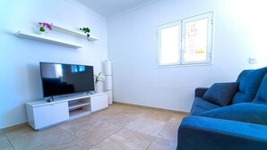 Una sala de estar con un televisor de pantalla plana sobre un soporte blanco, un sofá azul y una ventana con persianas.