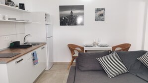 A compact living space with a kitchenette, a gray sofa with patterned pillows, a dining table with chairs, and two black-and-white cityscape photographs on the wall. - One bedroom apartment (TAHICHE)