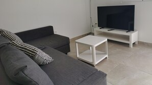 A living room with a grey sofa, a white coffee table, a flat-screen TV on a white stand, and a painting of a beach scene on the wall. - One bedroom apartment (TAHICHE)