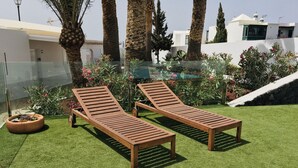 Two wooden lounge chairs on artificial grass, surrounded by palm trees and flowering plants, with a white building in the background. - One bedroom apartment with terrace. (TAHICHE)