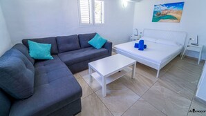 A modern living room with a grey sofa, a white coffee table, and a bed with a white headboard. There is a painting on the wall and a window with blinds. - One bedroom double apartment whit terrace (TAHICHE)