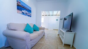 A living room with a beige sofa, teal cushions, a flat-screen TV on a white stand, a painting of a coastal scene, and a tiled floor. - One bedroom apartment with pool views (TAHICHE)
