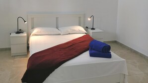 Cama bem arrumada com lençóis brancos, manta vermelha e toalha azul. Há duas mesas de cabeceira com luminárias de cada lado da cama.