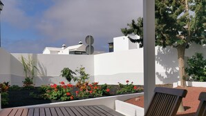 Um pátio com mesa e cadeiras de madeira, cercado por um jardim com flores vermelhas e uma parede branca ao fundo.