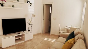 A living room with a flat-screen TV on a white entertainment unit, a beige sofa with colorful cushions, and a small table with a lamp.