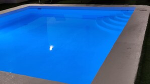 Une piscine rectangulaire aux parois bleues, entourée de chaises longues et d’un mur en briques.