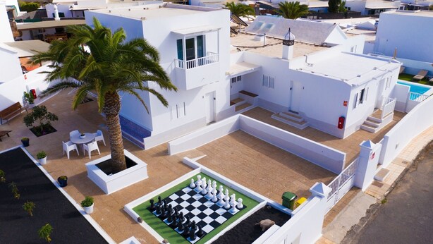 Un bâtiment moderne de couleur blanche, doté d’une terrasse sur le toit où l’on trouve un échiquier et des palmiers.