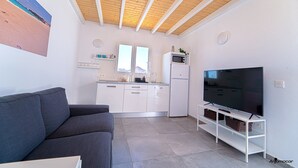 Un salon moderne avec un canapé gris, un téléviseur à écran plat posé sur un meuble blanc, un coin cuisine doté de meubles blancs et d’un four à micro-ondes, et un plafond en bois.