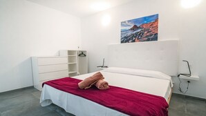 Una camera d'albergo con un letto, una cassettiera bianca, un asciugamano rosso sul letto, un quadro al muro e una mensola bianca.