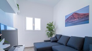 Ein modernes Wohnzimmer mit einem grauen Ecksofa, einem Flachbildfernseher, einem kleinen Tisch mit Pflanze und einem Fenster mit Jalousien.