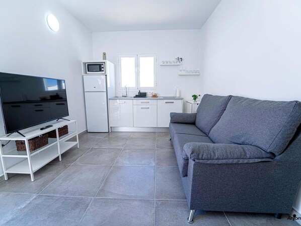 Una sala de estar moderna con un televisor de pantalla plana, un sofá gris, muebles blancos y una pequeña cocina.